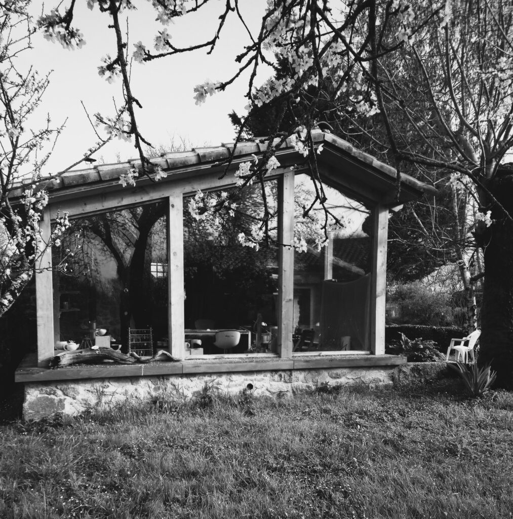 photo noir et blanc prise au primtemp de l'atelier Terre de Djen. Structure entièrement vitrée sur un sous bassement pierre. Avec en premier plan une vu sur des branches de prunus en fleurs