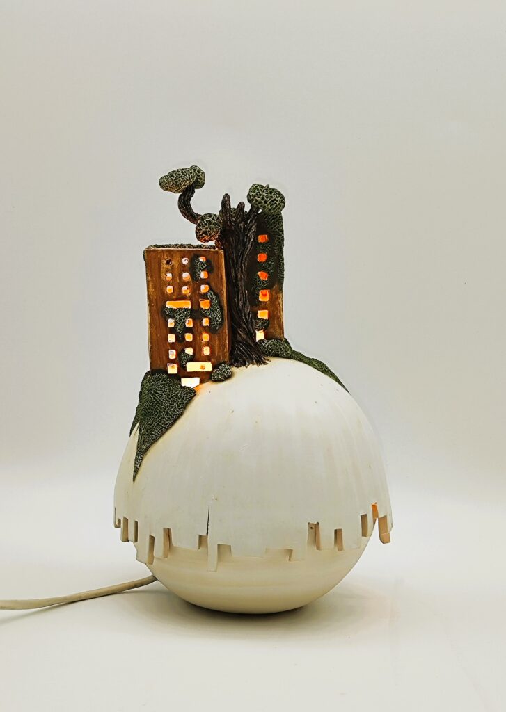 Ce luminaire aux formes ronde scindé en deux parties représente une cité miniature surgissant d’une colline, envahie par une végétation onirique. La lumière s’échappe des ouvertures comme des fenêtres habitées, diffusant une lueur douce qui transforme l’ensemble en un fragment de monde suspendu entre conte et rêve.