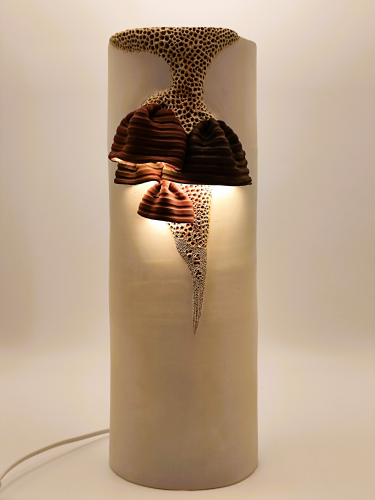 Ce luminaire, aux lignes élancées et organiques. Modelé en grès blanc laisse émerger des formes texturées imitant presque les champignons semblant se déployer le long de sa surface. La lumière s’y diffuse avec douceur, révélant les reliefs créant une présence chaleureuse et vivante.