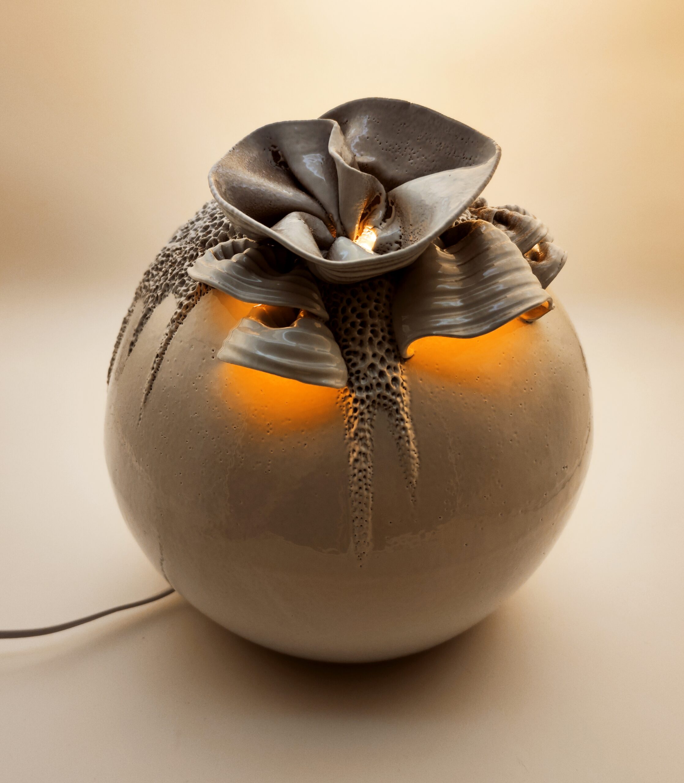 Luminaire inspiré du monde discret et poétique des champignons. Modelé en argile marron, il évoque une forme organique en pleine éclosion. L’émail blanc souligne ses reliefs et révèle, lorsque la lumière s’allume, une lueur douce et chaleureuse semblant naître de l’intérieur.